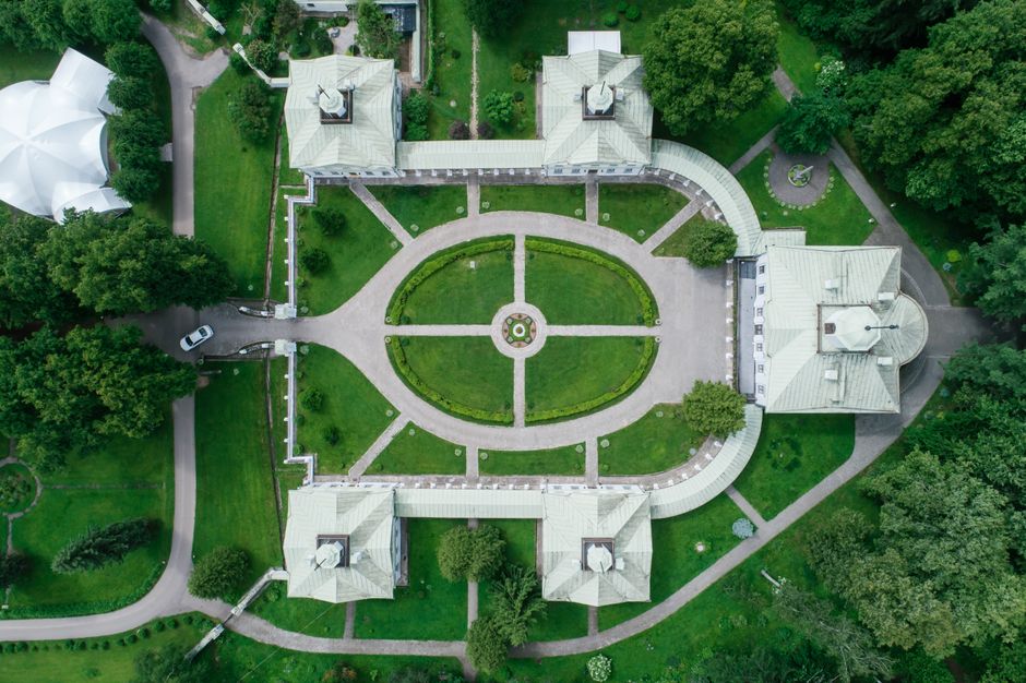 An aerial view of Avery Hill Park showcases a symmetrical formal garden enclosed by a white fence, with manicured lawns divided into geometric sections by paved pathways. The central lawn features a d