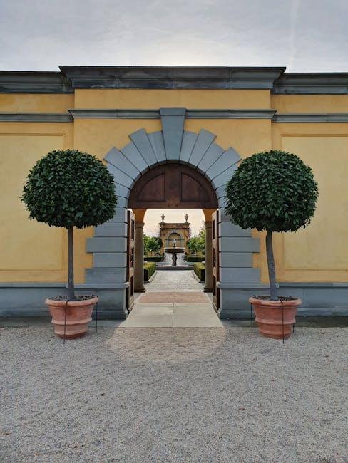 A curved gravel pathway winds through a well-maintained garden adjacent to a series of glass greenhouses with sloped, transparent roofing supported by white metal frames. The garden beds along the pat
